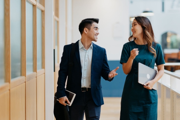An Asian man wearing a dark suit holds a book while walking through an office talking to an Asian female colleague with long dark hair and blue dress. She holds a laptop in her left hand